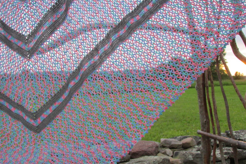 completed summer wildflowers triangle shawl in front of grassy hillside backround, stretched to see stripe pattern clearly.
